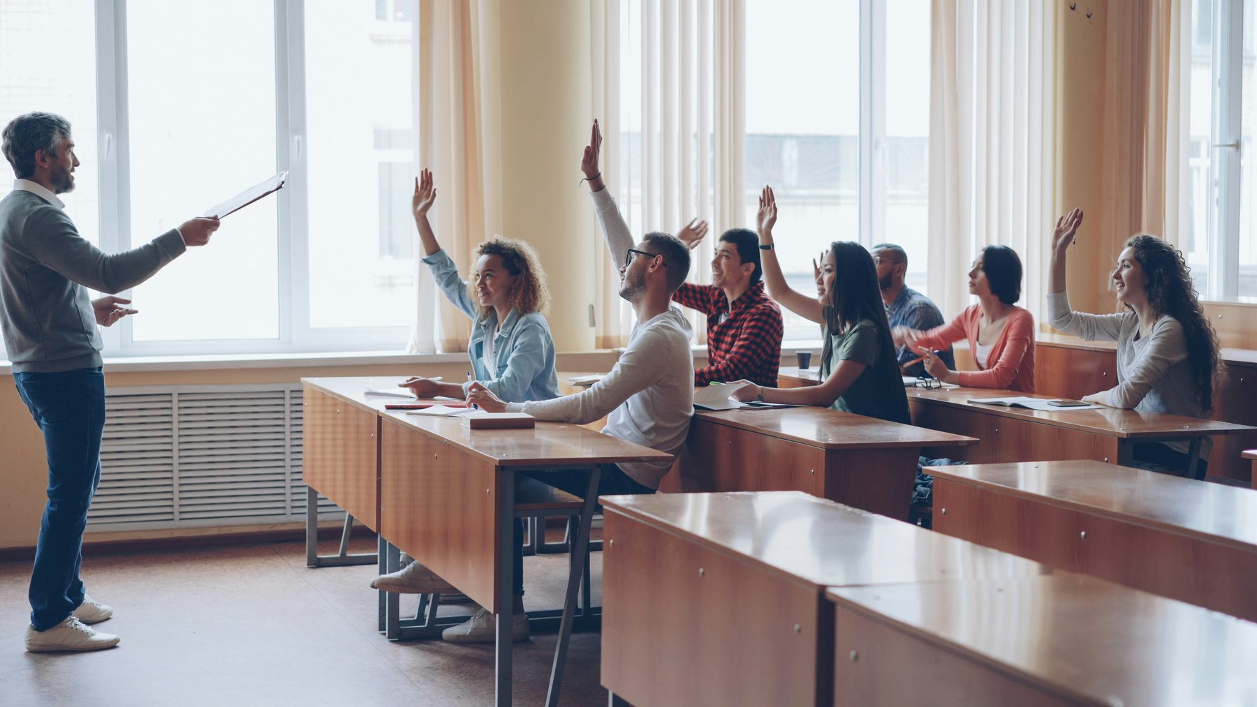 Students in a classroom raising their hands while a teacher stands at the front.